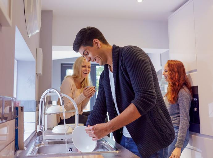 A group of happy college students laughing in the kitchen while one friend washes the dishes