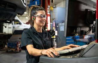 A women in an automotive shop searches for a part number on a computer, with a vehicle in the background.