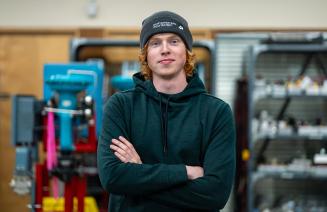 Student wearing a toque posing with arms folded in a workshop with machinery and tools in the background