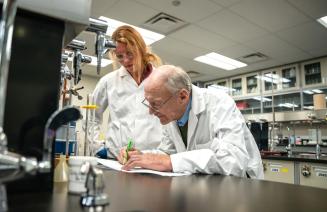 A student and organic chemistry instructor in lab coats collaborate on paperwork at a desk, focused on their research and findings