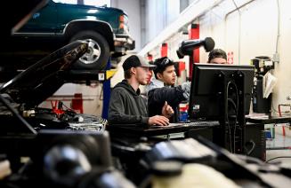 3 men collaborating around a computer in an auto repair shop surrounded by tools and and a lifted vehicle