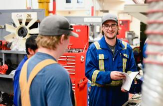 Two men in blue overalls chatting in a workshop with tools in the background.