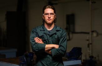 Tradesperson wearing protective eye wear and coveralls, standing in a workshop with arms folded.