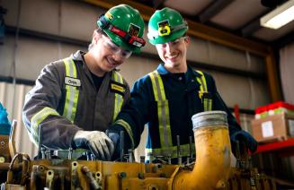 Two tradesmen wearing coveralls and hard hats share a laugh while working on equipment