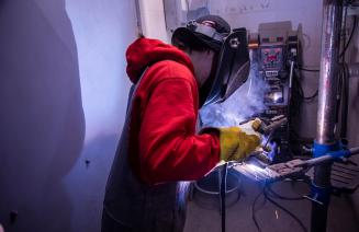 A person equipped with a welding helmet and gloves is engaged in welding, surrounded by bright sparks and metal.