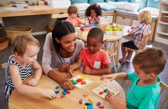 An happy early childhood educator in a classroom engaging with young children sitting around a crafting table playing a game.