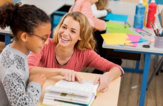 A teacher crouched beside a child sitting at a desk and helping her to read