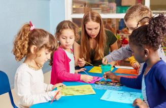 A teacher interacting with a group of children who are drawing pictures on a crafting table
