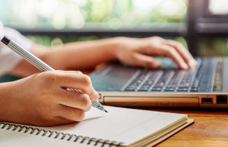 Student researching on a laptop while writing notes in a notebook