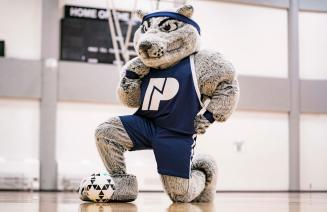 Farley the mascot kneeling in the gym with a soccer ball at his feet