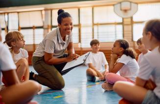 Phys Ed teacher smiling and pointing to a clipboard while addressing a group of school children in the gymnasium