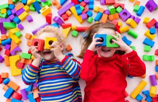 Two happy children laying down and holding building blocks up to their eyes and surrounded by lots of colourful blocks