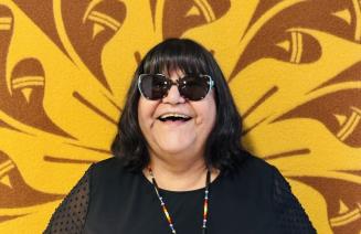 Portrait of Elder Theresa happily smiling in front of the Indigenous blanket that hangs outside the on-campus friendship centre