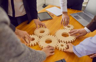 Company workers standing around a table joining cogwheels as metaphor for teamwork, business system and working solutions