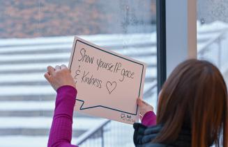 Student holding up a poster with a handwritten phrase inside a speech bubble that says "show yourself grace and kindness."