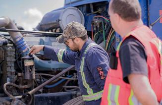 A driving instructor observes a student checking the engine of a semi-truck.