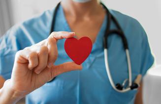 A nurse wearing blue scrubs with a stethoscope around her neck holding up a red heart between her thumb and index finger