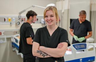 A happy practical nursing student in the simulation lab, posing with arm folded as fellow students work in the background.