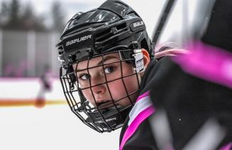 A female hockey player wearing a helmet looking directly at the camera from the team bench