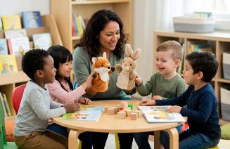 A smiling female teacher engages four diverse young children in a puppet show activity at a small round table in a bright classroom. 