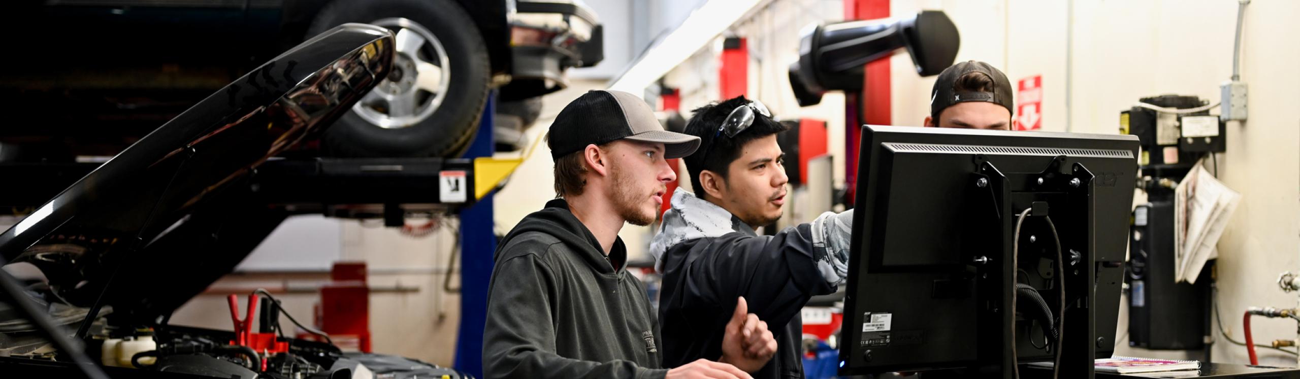 3 men collaborating around a computer in an auto repair shop surrounded by tools and and a lifted vehicle