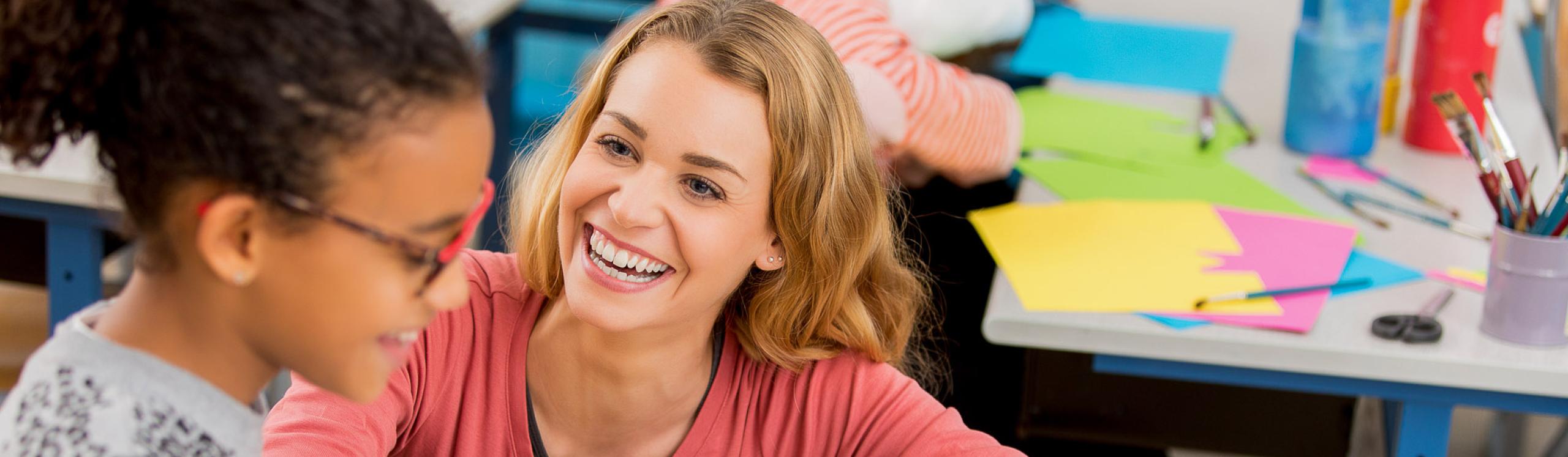 A teacher crouched beside a child sitting at a desk and helping her to read