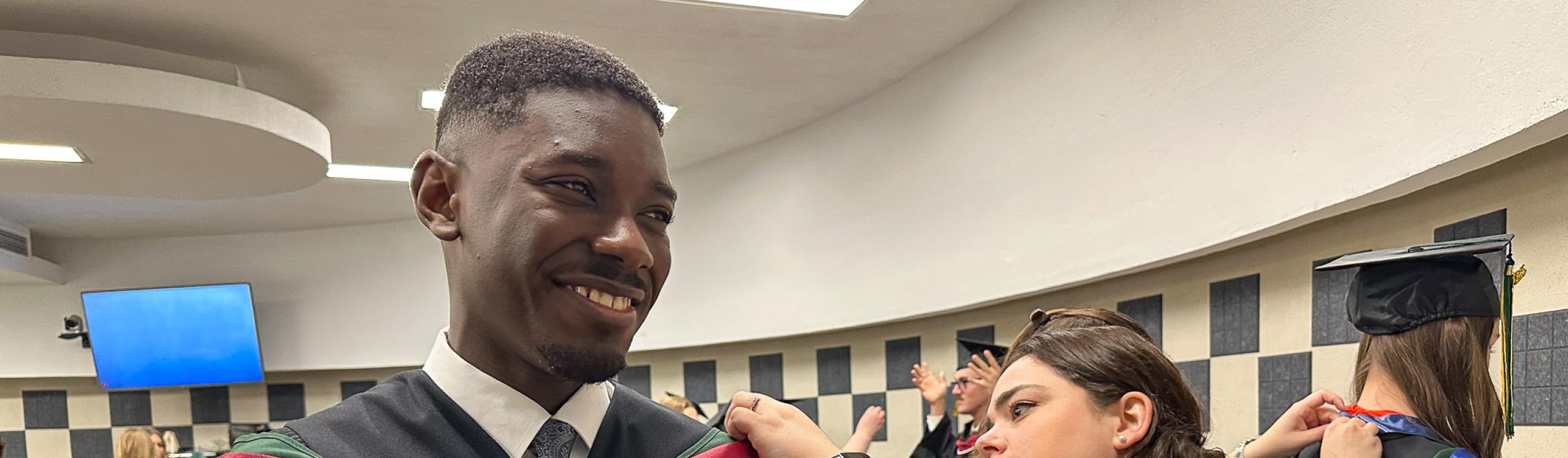 2025 Valedictorian Clinton Owusu smiles while receiving assistance gowning up before the Convocation ceremony