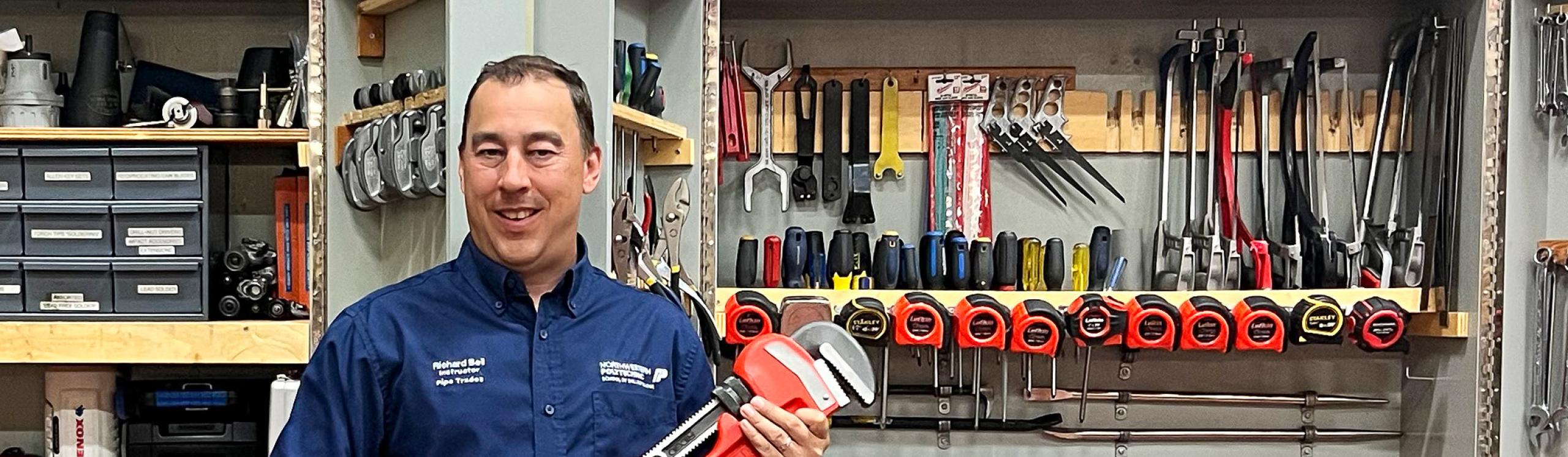 NWP plumbing and gasfitter instructor, Richard Bell holding a large wrench, posing in front of a tool cupboard.