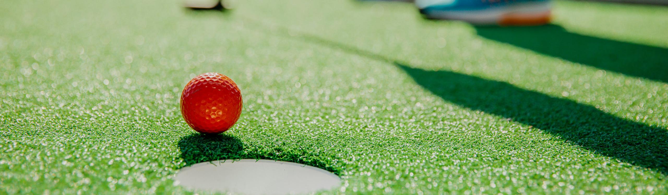 a red golf ball right beside the hole on a mini golf course with the feet and putter of the golfer in the background