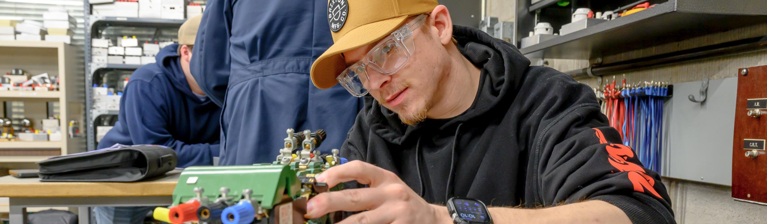 An electrician student wearing safety googles closely observing a piece of electrical equipment in the electronics lab at NWP