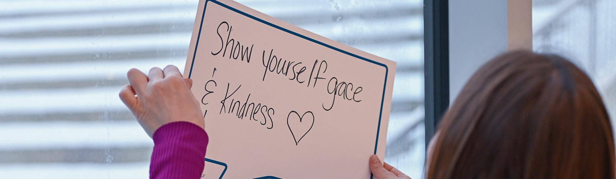 Student holding up a poster with a handwritten phrase inside a speech bubble that says "show yourself grace and kindness."
