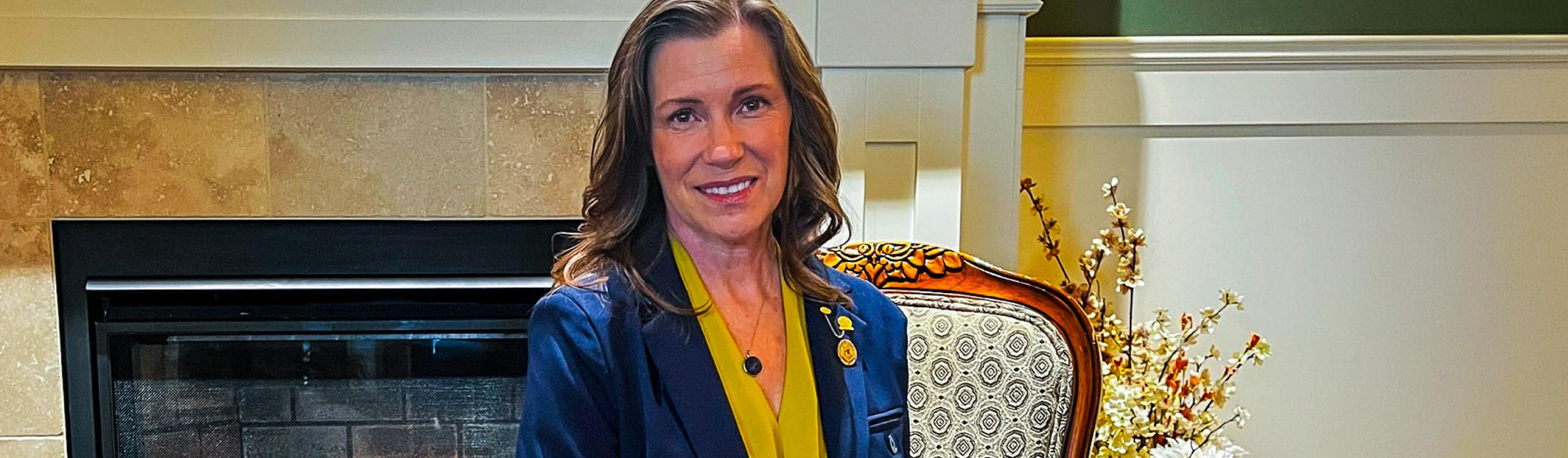Smartly dressed NWP Alumni, Eunice Friesen seated on an ornate chair in front of a warm and inviting fireplace