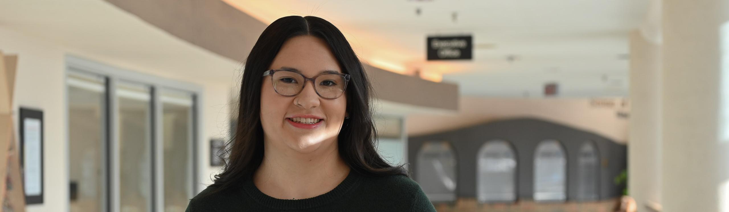 Jai Lin Mark, NWP alumna, posing in the E-wing corridor at NWP with warm sunshine streaming through the windows behind her
