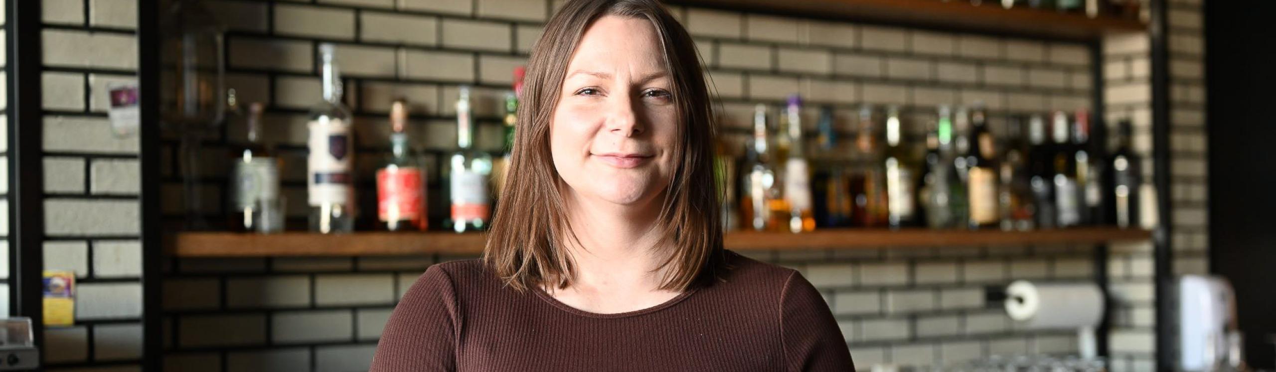 Jessica Schoepp, 92 Beverage Co owner and NWP Alumna standing behind the bar with an engaging collection of liquor in the background