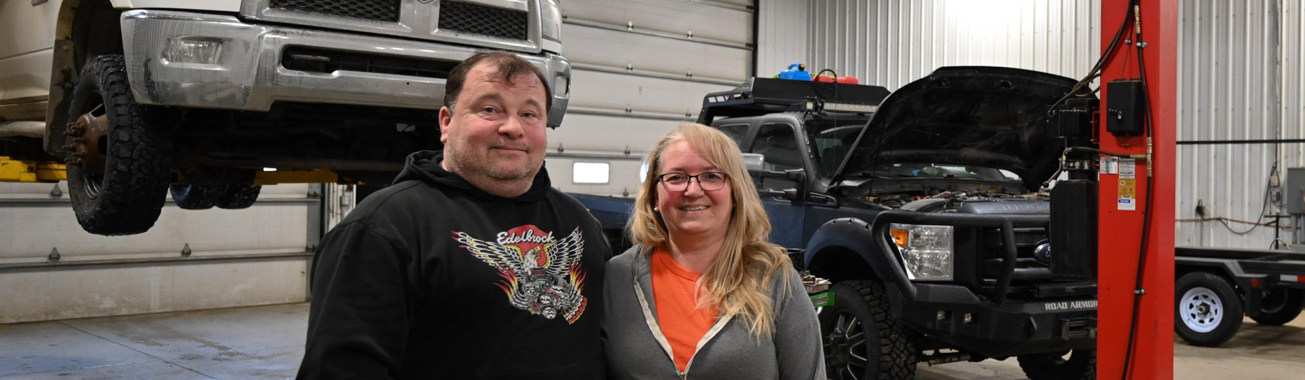 Danielle and Stacey Commandeur stand together inside their automotive and heavy-duty repair shop, with two pickup trucks behind them in the service area.