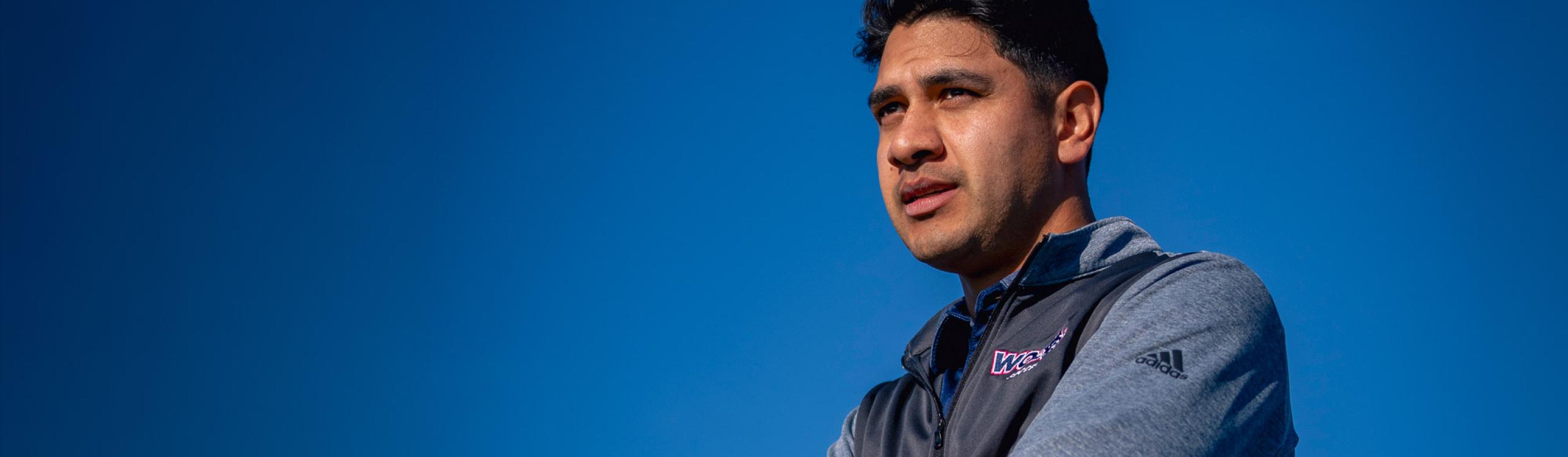 Mens soccer head coach, David Rojas observing the team play with a bright, clear blue sky in the background