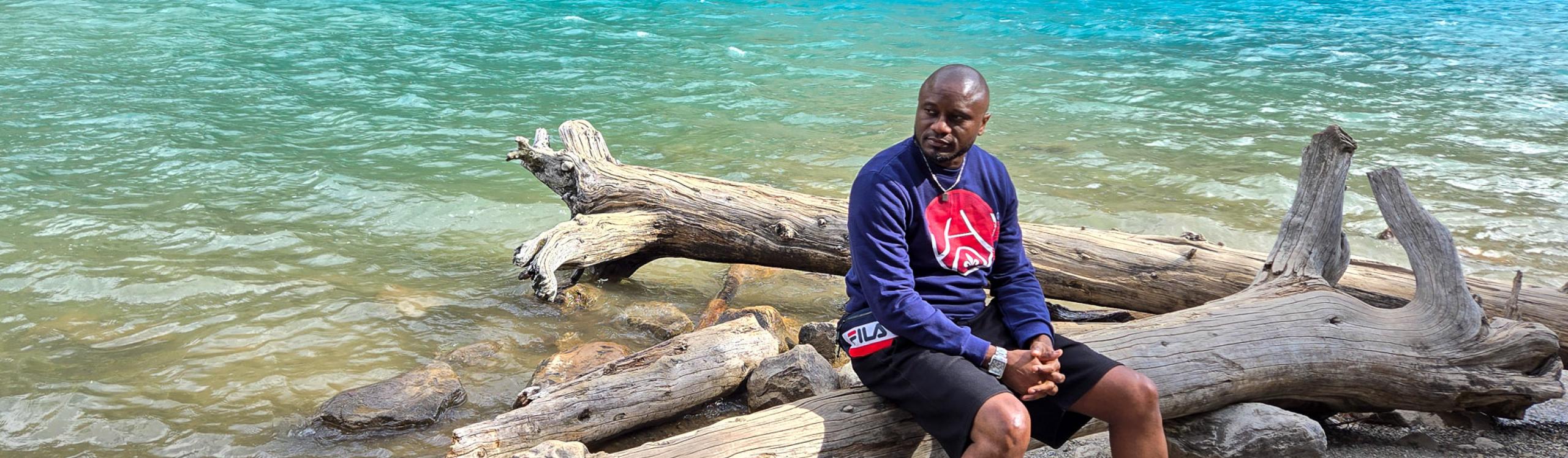 Phil Mvondo seated on a large tree trunk near an aquamarine-colored lake, surrounded by the stunning landscape of the Canadian Rockies.